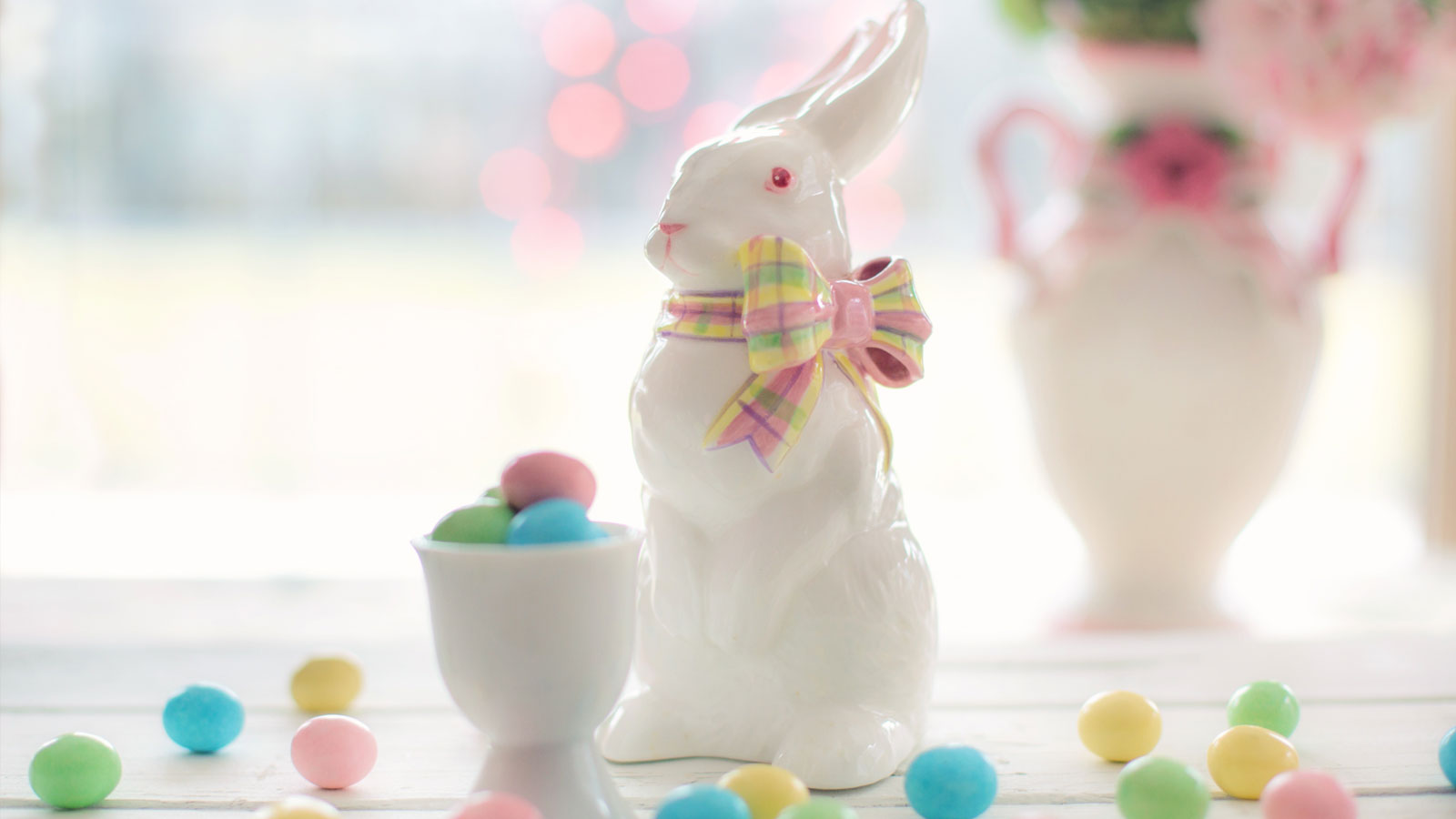 Bunny figurine with plaid bow with pastel Easter candy in an egg cup and on the table